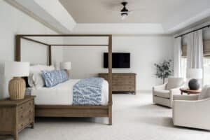 Comfortable bedroom with four post bed, seating area with two swivel chairs in a beige fabric, large table lamps, custom linen curtains with nature woven shades. Paint includes white walls with a greige contrast trim and ceiling above tray is painted same color.