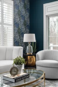 Seating area with grey swivel chair, white velvet sofa, glass lamp with gold accents sitting on a black side table. The coffee table is glass with gold metal finish. Beautiful floral wallpaper paired with a teal paint color.