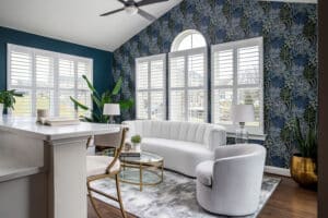 Sunroom with beautiful floral wallpaper that is paired with a teal wall color. Modern curve of the white velvet sofa paired with grey swivel chair. Lamps and coffee table have gold metal and glass. Modern black ceiling fan.