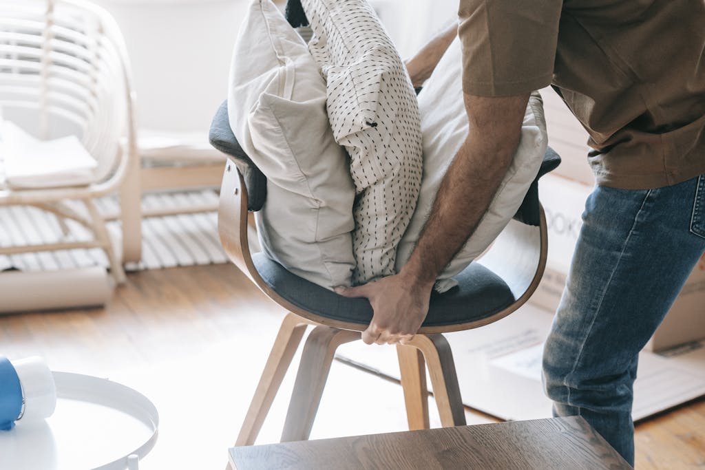 A man packing throw pillows on a contemporary chair indoors, suggesting moving or organizing.