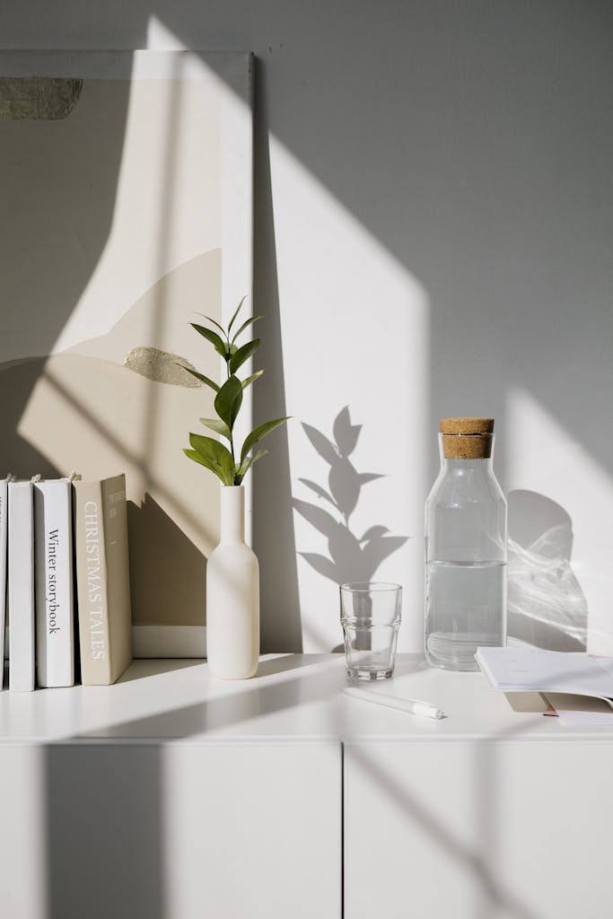 A modern minimalist setup featuring books, a vase, and a carafe on a sunlit shelf.