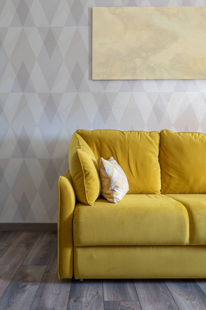 Bright yellow sofa with decorative cushions in a modern living room featuring geometric wallpaper and wooden flooring.