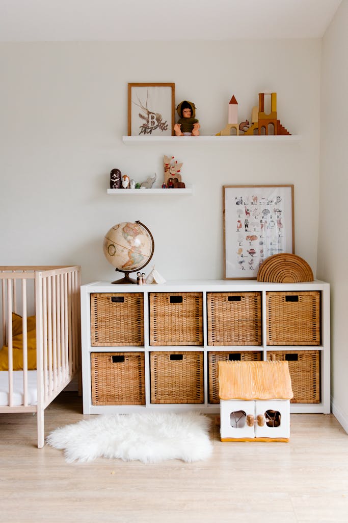 Comfortable bed with toys globe and paintings placed on wooden cabinet with wicker boxes and soft white rug with toy house on floor