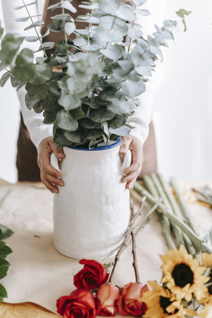 Crop anonymous florist standing with stems in vase and roses with sunflowers on table